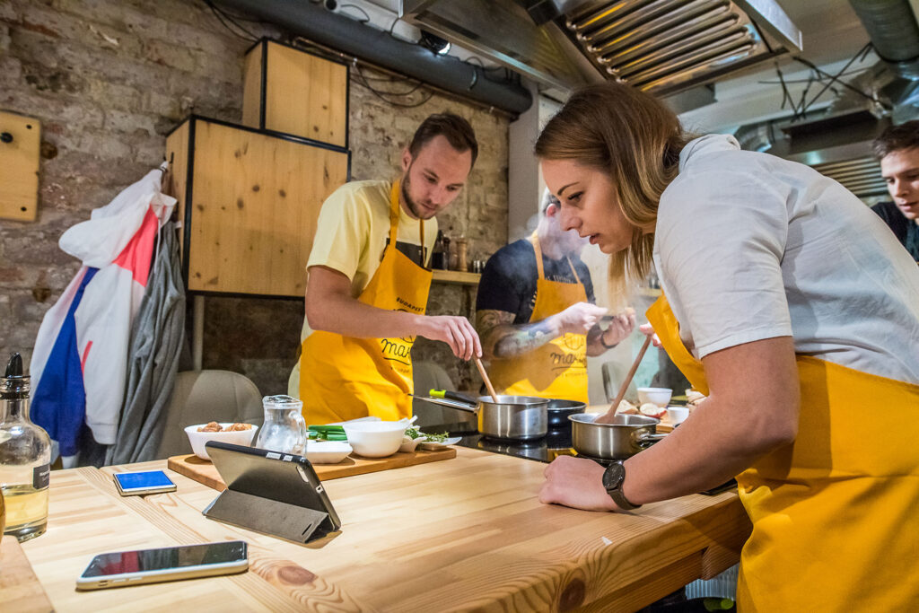 főzőiskola, egy nő és egy férfi sárga kötényben áll egy asztalnál, kezükben fakanál
