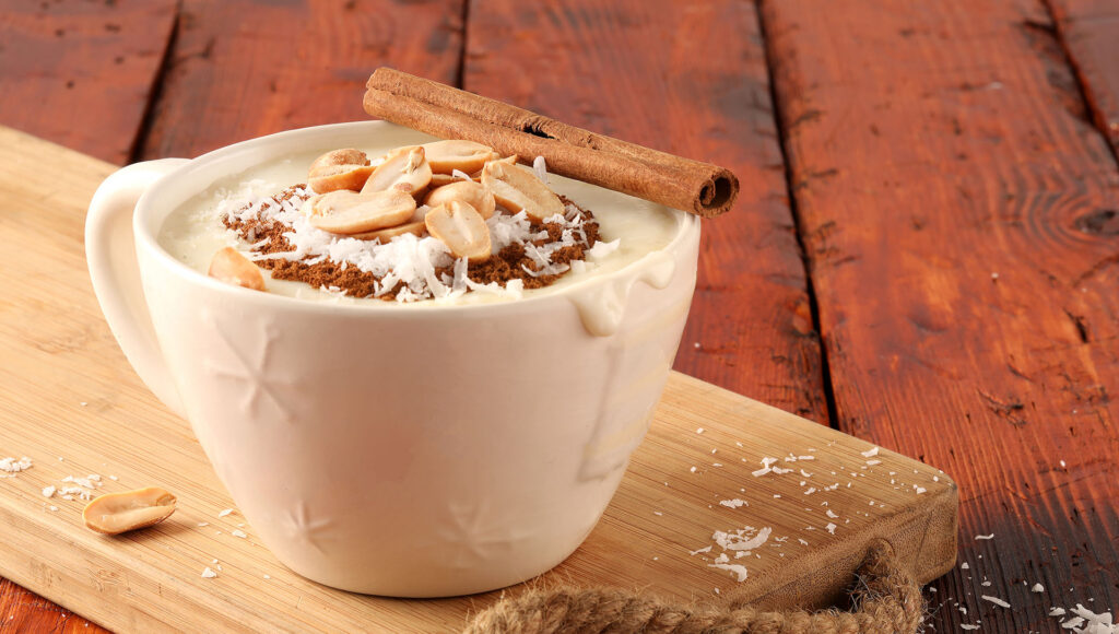 latte, fehér csészében barna ital, rajta barna fahéjrúd és mogyoró