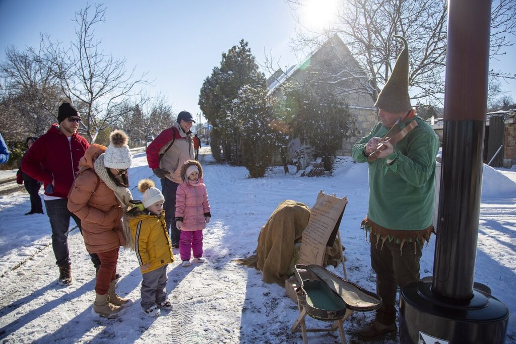 hóban gyerekek állnak szüleikkel, a kép jobb oldalán egy manónak öltözött ember hegedül
