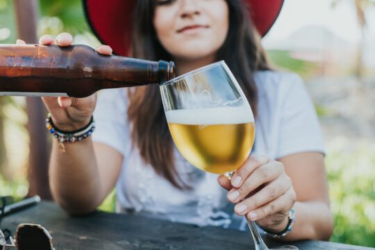 egy kalapos nő barna üveget és átlátszó poharat tart a kezében, utóbbiban sör