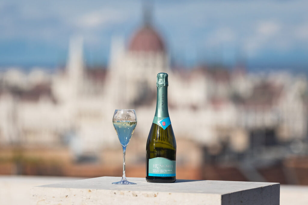 kék-barna hungaria pezsgő palack, mellette egy talpas pohár, mögötte egy épület