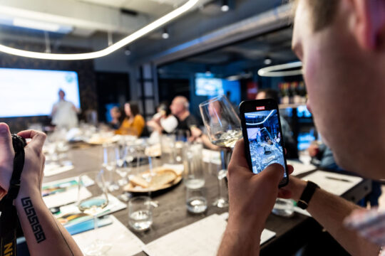 borkóstoló, egy férfi mobiltelefont és poharat tart a kezében, asztal körül emberek