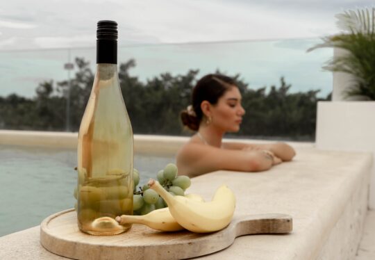 egy nő egy borhotel medencéjében relaxál, előtte egy vágódeszkán déli gyümölcsök és egy palack bor
