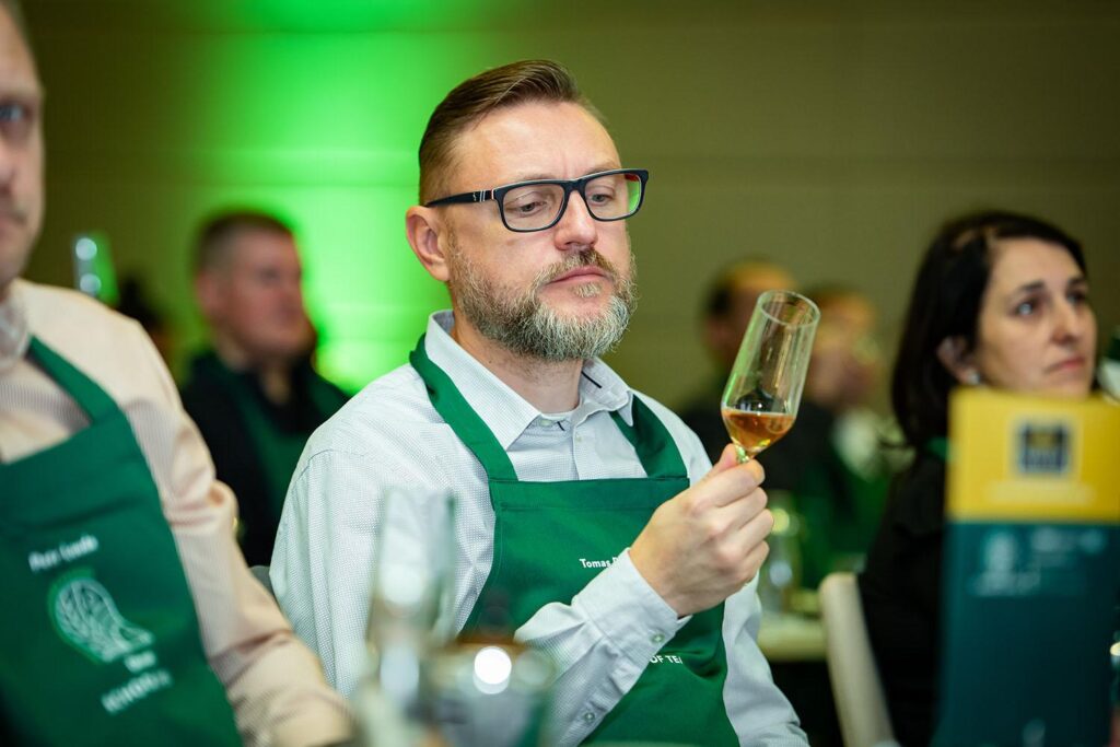 zöld kötényes szemüveges ember poharat tart a kezében, benne tea